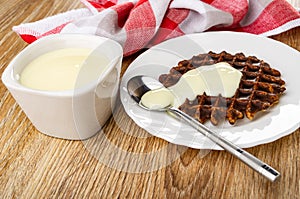Napkin, bowl with condensed milk, spoon with milk and wafer in chocolate on white plate on wooden table
