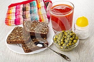 Napkin, bread with sunflower seeds in plate, spoon, glass with tomato juice, salt, bowl with green peas on wooden table