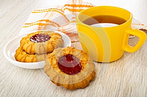 Napkin, cookies with jam in saucer, yellow cup with tea on wooden table