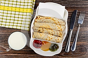Napkin, cup of milk, dish with jams, pancake rolls, tissue, fork and knife on wooden table. Top view