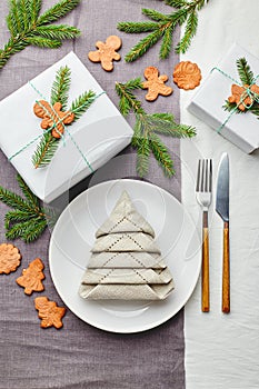 Napkin in the form of a Christmas tree on a plate on white tablecloth with gifts and decorations with fir sprigs and gingerbread