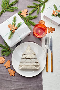 Napkin in the form of a Christmas tree on a plate on white tablecloth with gifts and decorations with fir sprigs and gingerbread