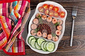 Napkin, partitioned dish with fried sausages, red rice, cucumbers, tomato, fork on table. Top view