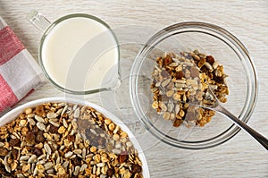 Napkin, pitcher with yogurt, muesli with cereals, raisin and sunflower seeds in plate, spoon and granola in bowl on wooden table.