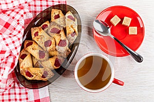 Napkin, shortbread cookies with jam in brown dish, cup with tea, sugar and teaspoon on saucer on table. Top view