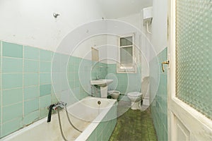 Narrow bathroom with green tiles and dirty floors, bathtub and white toilets