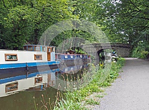 Boats and barges moored on the rochdale canal in hebden bridge bext to an old stone footbridge surrounded by green summer