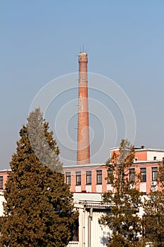 Narrow old red bricks tall industrial chimney with metal stairs mounted on side rising high above elongated factory building