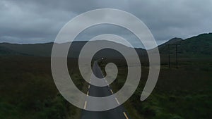 Narrow road with dashed lines marked edges in landscape at dusk. Fly against car arriving from distance. Ireland