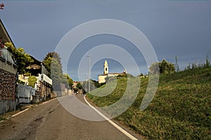 Narrow road on a hill bordered by an embankment with a viillage and a bell tower in the distance
