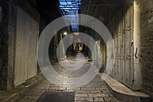 Narrow side street with closed white doors on each side and two dark human figures in the distance. Deserted urban road with stone