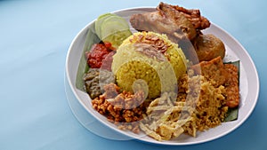 Nasi Kuning or Yellow rice with traditional fried chicken, tofu, tempeh, and smashed potato on blue background.