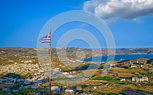 National flag of Greece waving over landscape of Milos island. Adamantas town, bay and villages at sunset. Whitewashed