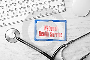 National health service (NHS). Badge with text, stethoscope and keyboard on white wooden background, flat lay