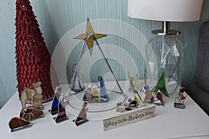 Nativity scene made of colored glass and metal frames on a small end table next to a lamp and stylized Christmas tree