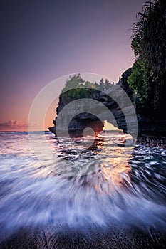 Natural arch. Batu Bolong temple on the rock during sunset. Seascape background. Motion milky waves on black sand beach. Copy