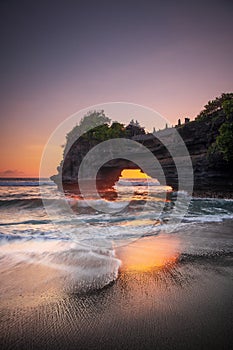 Natural arch. Batu Bolong temple on the rock during sunset. Seascape background. Motion milky waves on black sand beach. Copy