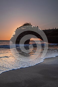 Natural arch. Batu Bolong temple on the rock during sunset. Seascape background. Motion milky waves on black sand beach. Copy