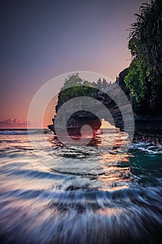 Natural arch. Batu Bolong temple on the rock during sunset. Seascape background. Motion milky waves on black sand beach. Copy