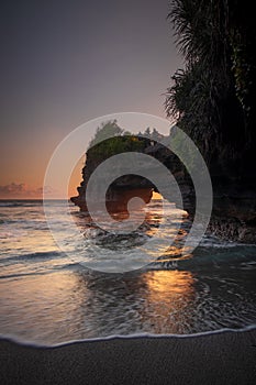 Natural arch. Batu Bolong temple on the rock during sunset. Seascape background. Motion milky waves on black sand beach. Copy