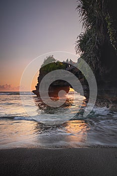 Natural arch. Batu Bolong temple on the rock during sunset. Seascape background. Motion milky waves on black sand beach. Copy