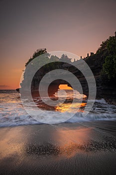 Natural arch. Batu Bolong temple on the rock during sunset. Seascape background. Motion milky waves on black sand beach. Copy