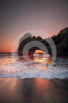 Natural arch. Batu Bolong temple on the rock during sunset. Seascape background. Motion milky waves on black sand beach. Copy