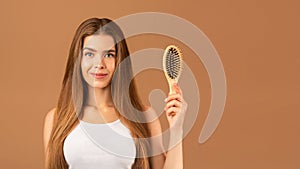 Natural beauty. Attractive young woman with perfect long hair holding wooden hairbrush on brown studio background