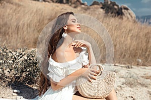 Natural beauty brunette outdoor portrait. Beautiful young woman  in a white dress with woven handbag enjoying in the hay field.