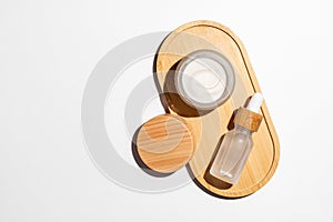 Natural cosmetics in white glass packaging on bamboo tray on white background. Open cream jar and dropper bottle with serum. SPA