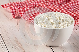 Natural cottage cheese in a white ceramic bowl with wooden spoon and red napkin on a wooden background. Diet concept