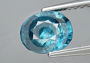 Natural gemstone blue zircon in tweezers on a gray background photo