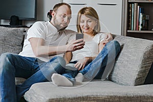 Natural, lifestyle image of attractive couple sitting on couch together looking at smartphone in the living room.