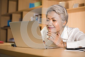 Natural lifestyle office portrait of attractive and happy successful mature Asian woman working at laptop computer desk smiling