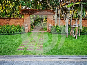 Natural orange clay brick arched wall entrance in a tropical garden with pattern of brown laterite walkway on green grass lawn