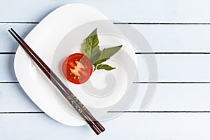 Natural organic tomato with green leaves and chopsticks on white plate for dietary breakfast or dinner. Top view on wooden table w
