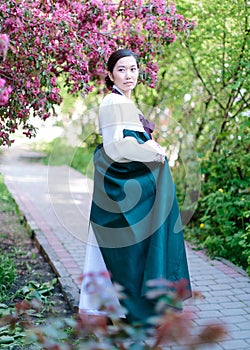 A natural portrait of a beautiful young Korean girl standing in the middle of a blossoming spring garden, holding the hem of a photo
