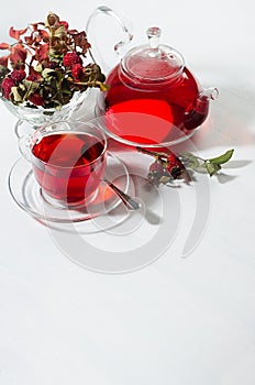 Natural red herbal tea in transparent teapot, cup with hawthorn berry, rosehip, dry leaves in morning sunlight on soft light white