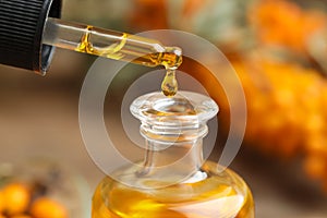 Natural sea buckthorn oil dripping from pipette into bottle on blurred background, closeup photo