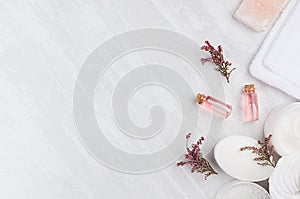 Natural spa cosmetics with pink rose oil, salt, soap and lavender flowers on white wood background, top view, border.