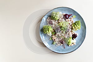 Natural vegan salad of mini beets, romanesco cabbage, white beans and sprouted onion seeds on a gray plate on a light background.