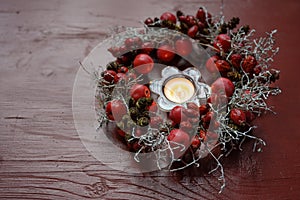 Natural wreath with rose hips and small apples around a burning candle on a red wooden table, seasonal decoration in autumn,