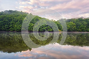 Tapeta přírodního zrcadlového světa, jemně růžový západ slunce nad řekou se zeleným kopcem a zataženou oblohou