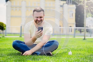Nature and technology. A man with a beard lies on a green grass with a smartphone in his hands is charging power bank.