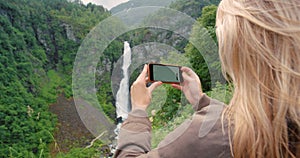 A nature tourist taking photos on phone of the green mountain, waterfall view on hiking adventure outdoors. Young