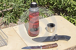 Nature and wooden table on which there is an old thermos, hunting knife, grill