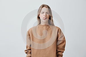 Naughty beautiful girl with blonde long hair in beige sweater misbehaving, sticking out tongue at camera as a sign of