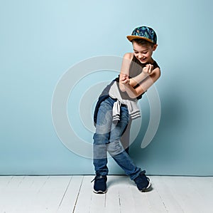 Naughty schoolboy in stylish cap jumping in place looking down under his feet with roguish smile. isolated on blue background