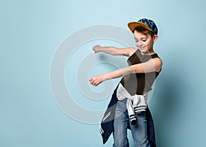 Naughty schoolboy in stylish cap jumping in place looking down under his feet with roguish smile.  isolated on blue background