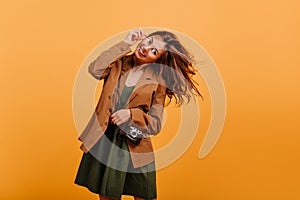 Naughty teen girl is having fun and posing on orange background. Student with camera takes off glasses and plays hair.